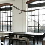 Ventilateur suspendu au-dessus d’une table dans une pièce lumineuse, grandes fenêtres et ambiance moderne.