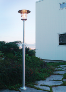 Luminaire bord de mer extérieur: lampe extérieure en inox 316 sur pied dans un jardin près d’une façade, émettant une lumière chaude.