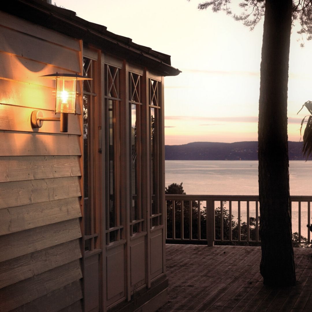 Applique murale lumineuse près d'une terrasse en bois, éclairant une façade en bois et une porte-fenêtre, avec vue sur l'eau au coucher du soleil. Comment choisir luminaire exterieur.