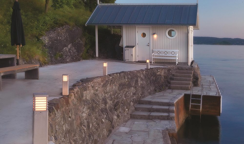 Allée pavée menant à une petite cabine blanche au bord de l’eau, avec des luminaires extérieurs allumés le long du chemin.