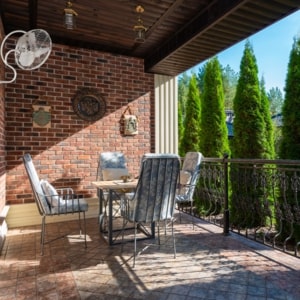 Balcon avec table et chaises autour, sous une terrasse couverte et un ventilateur mural sur le mur en briques.
