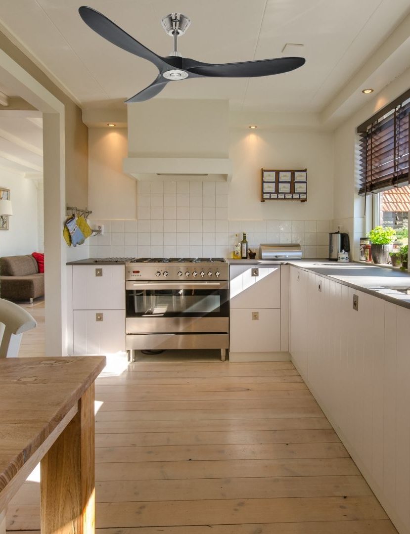 Cuisine lumineuse avec îlot et grand ventilateur mural au plafond, près d’une fenêtre ouverte et d’un plan de travail blanc.