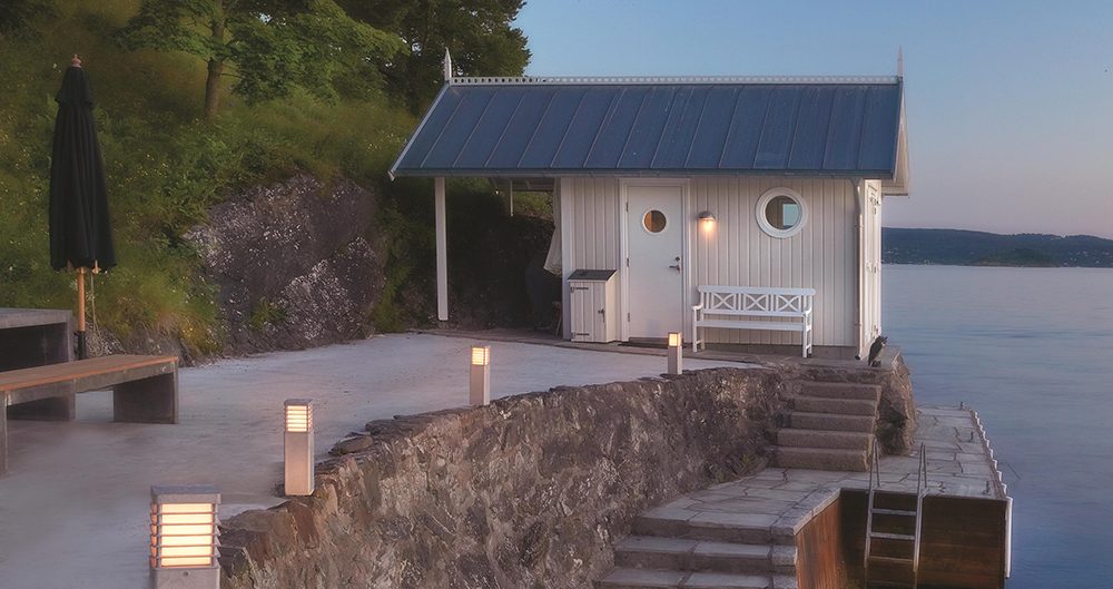 Éclairage extérieur devant une petite maison au bord de l’eau avec des poteaux lumineux le long d’une terrasse rocheuse.