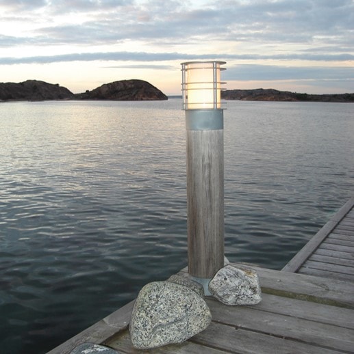 Luminaire extérieur en aluminium et bois sur une jetée, près de l’eau, avec une lampe cylindrique lumineuse en haut.