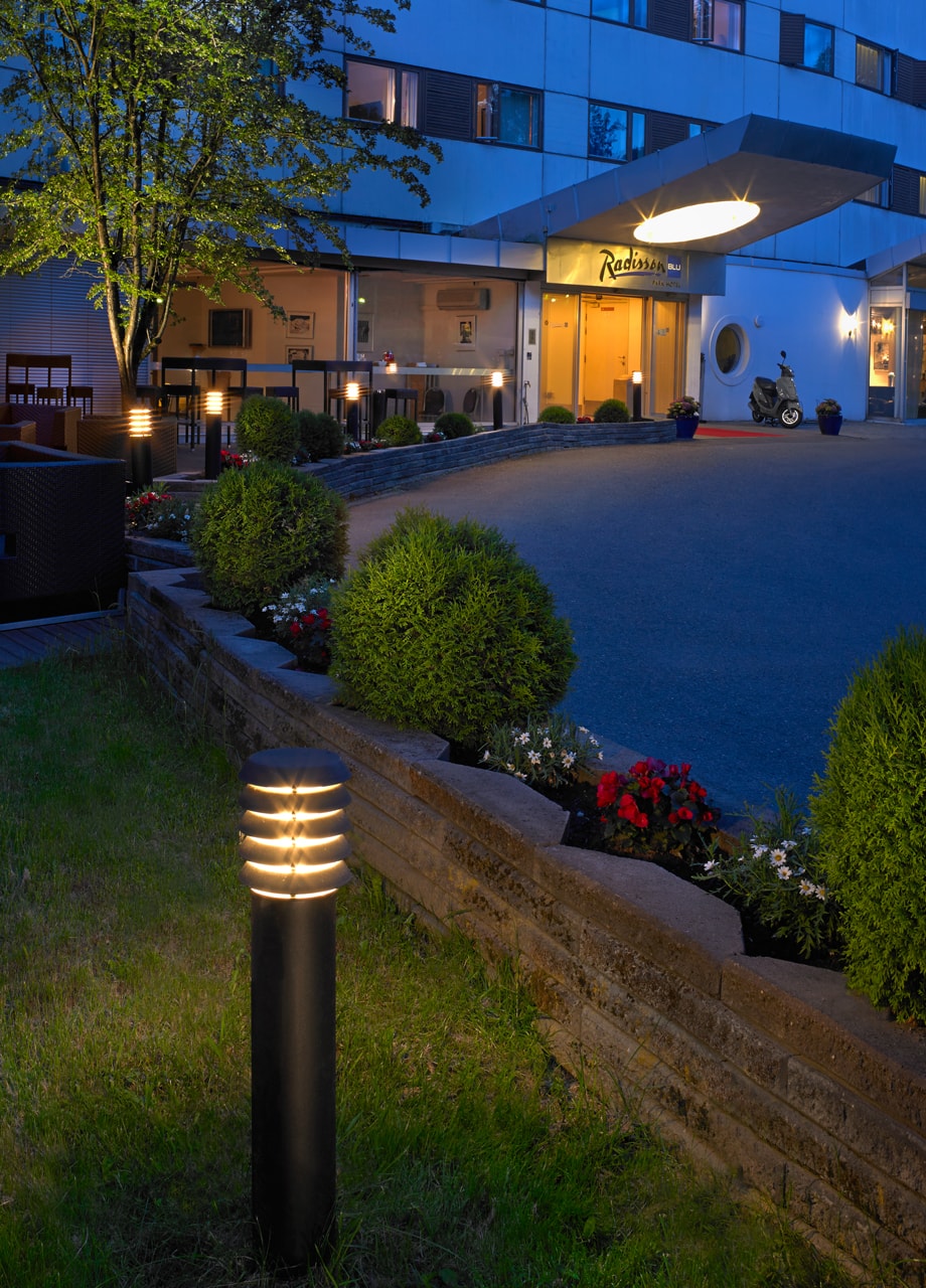 Luminaire de jardin en premier plan émettant une lumière chaude dans un espace extérieur près d’un bâtiment.