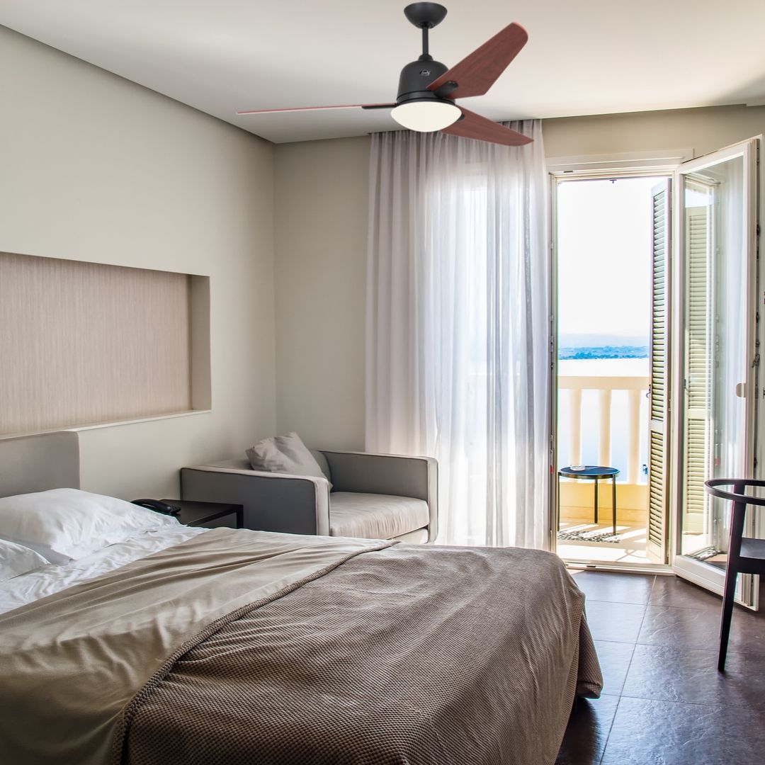 Ventilateur de plafond avec lumière allumé dans une chambre, près d’un balcon ouvert et d’un fauteuil gris.